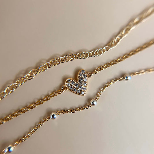 Flat lay of a delicate gold vermeil bracelet stack featuring a pavé heart charm, styled against a neutral background to highlight the charm’s sparkle and refined texture