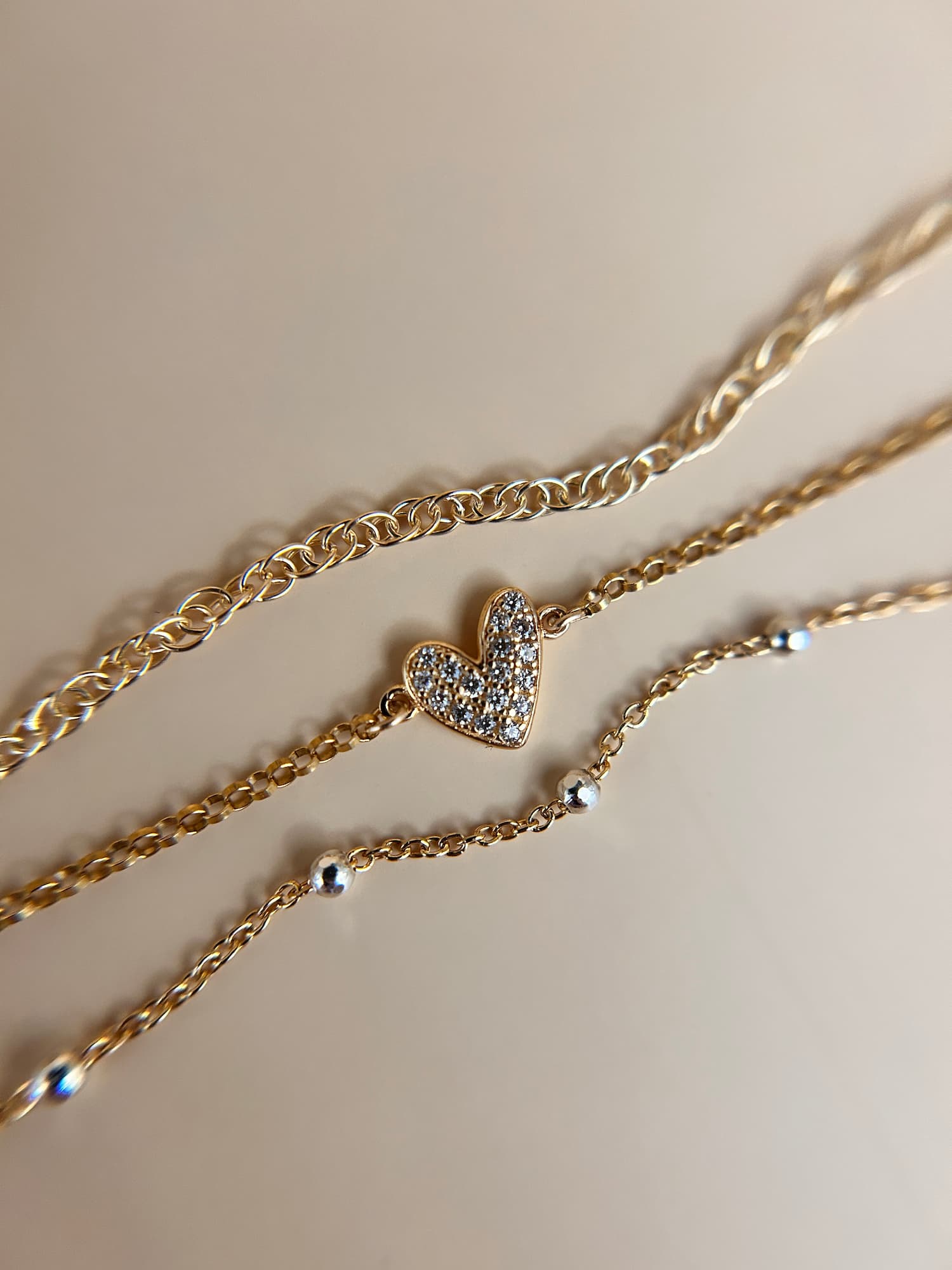 A close-up of layered gold-tone necklaces on a warm beige background, featuring a heart-shaped pendant covered in small clear stones and delicate chain details.