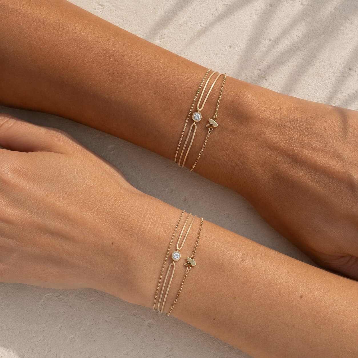 Two delicate gold-toned double-strand bracelets with a small round clear stone and tiny charm details, shown worn on two wrists against a light neutral textured background.